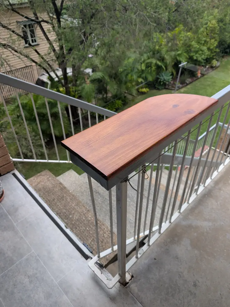 Stained timber drinks ledge with a rounded end, fitted to the top of a white balcony railing