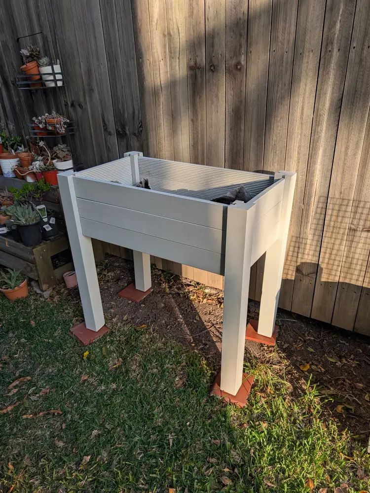 Elevated timber planter box on legs, painted pale grey, standing on pavers against a timber fence